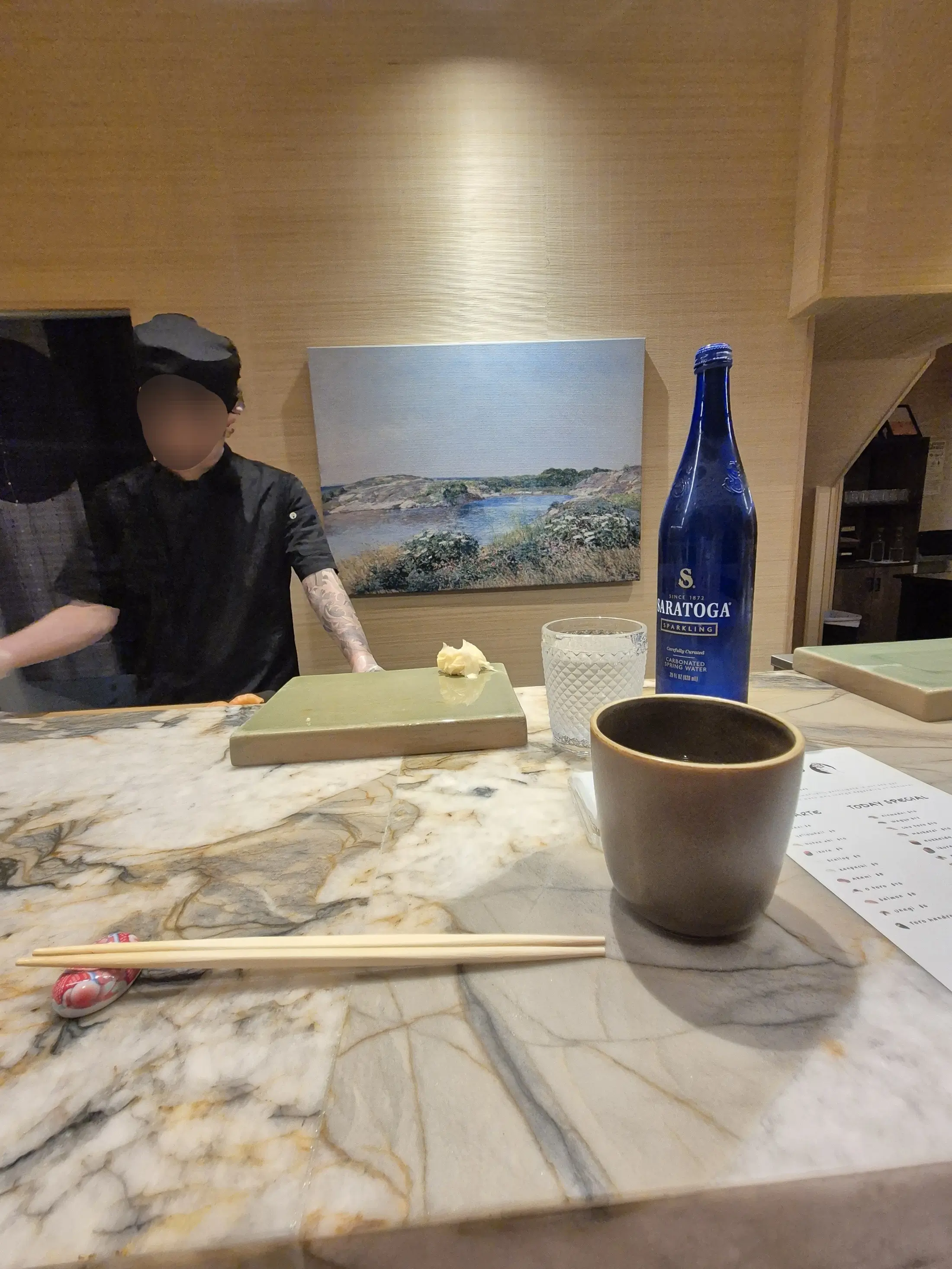 A tattooed chef in black stands behind a marble counter set with chopsticks and a blue water bottle.
