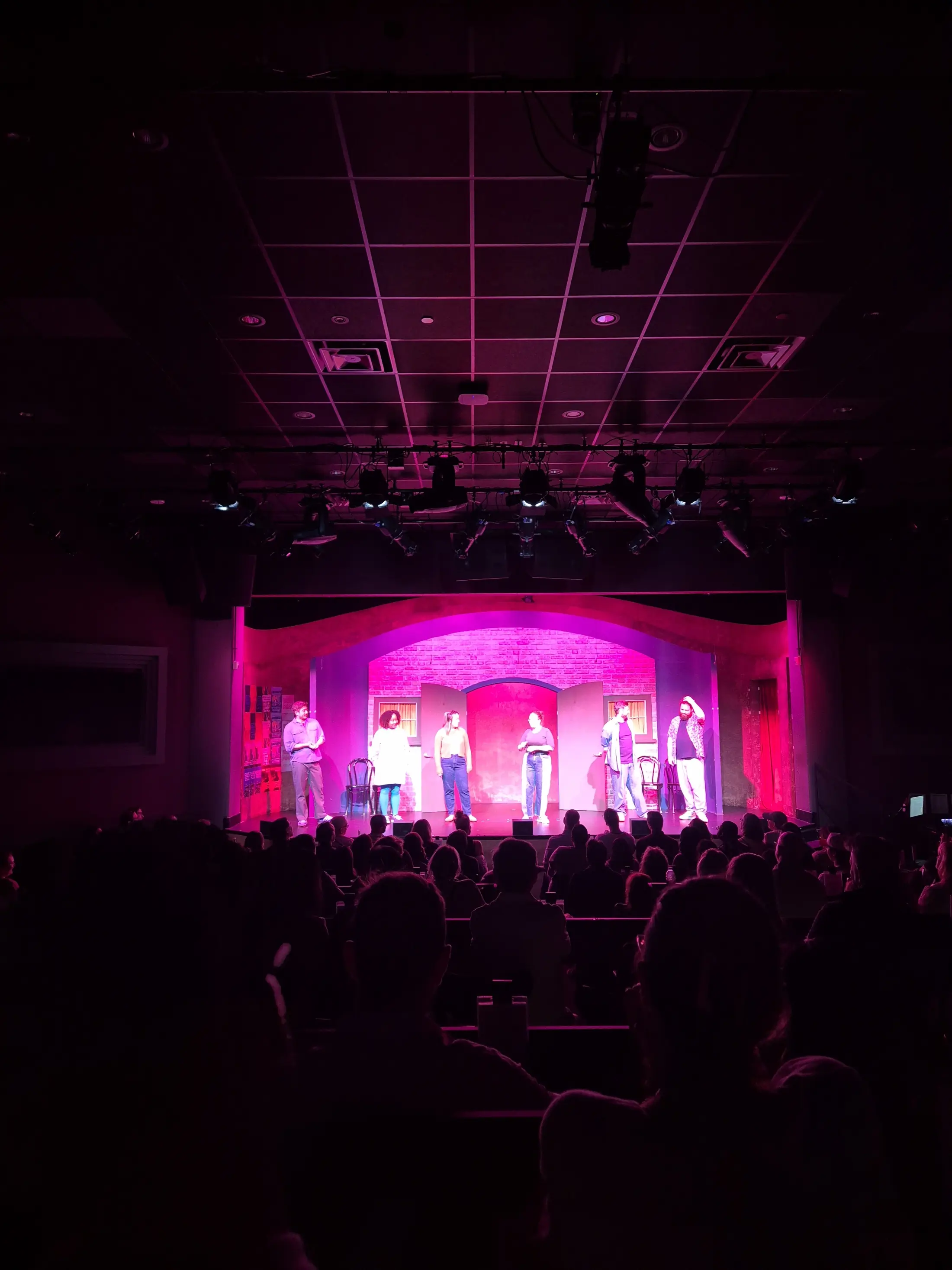 A lively theater scene with six performers on stage, bathed in dramatic pink light.