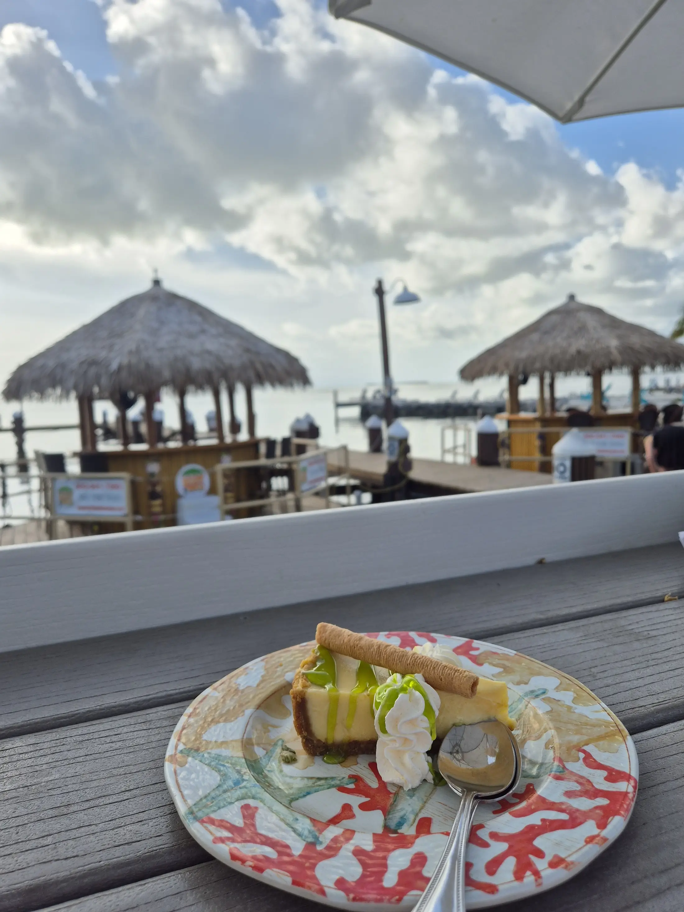 A slice of cheesecake with whipped cream by a seaside pier and thatched huts.
