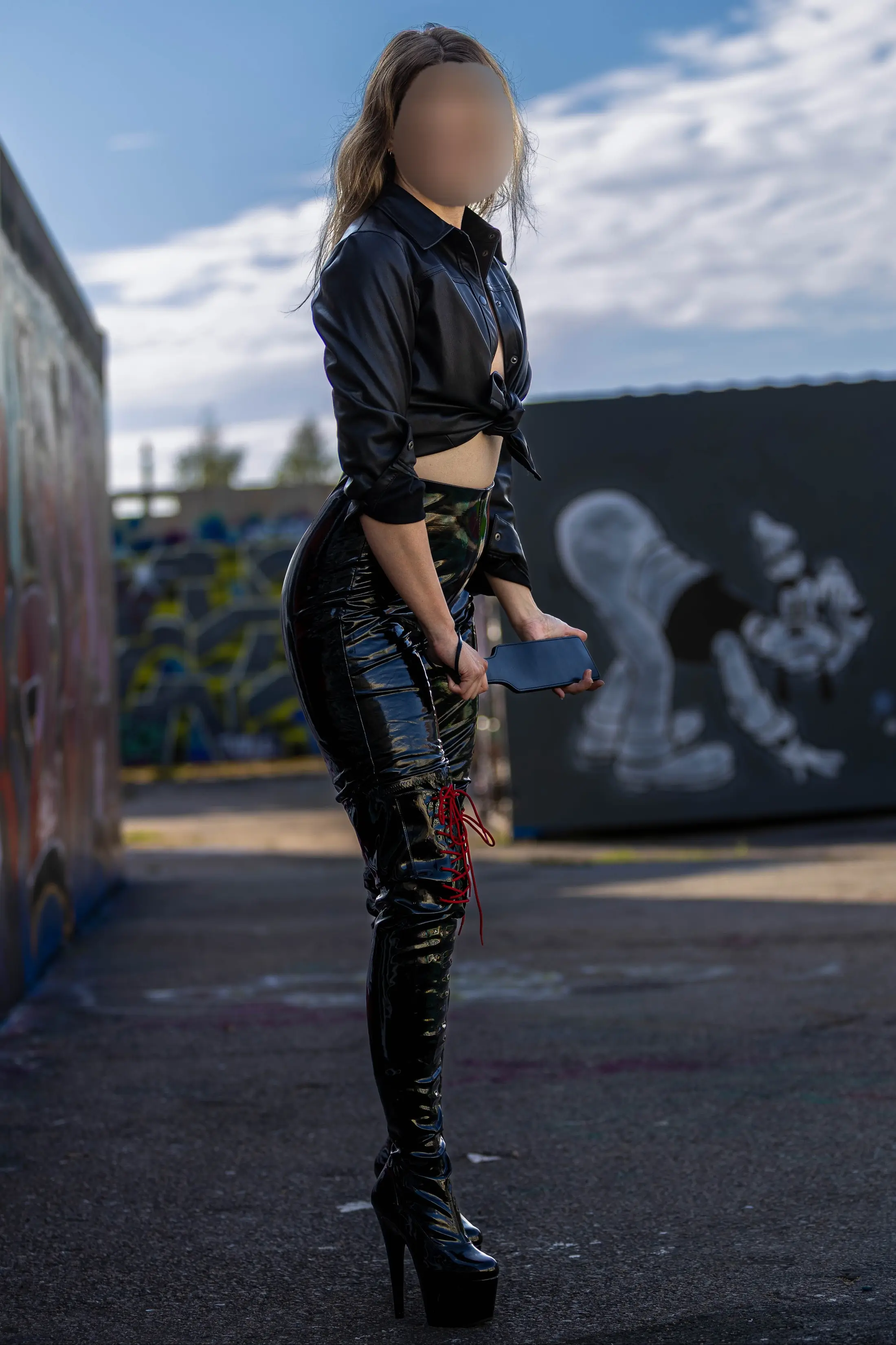 Josefine in sleek black leather and thigh-high boots stands by urban graffiti art.