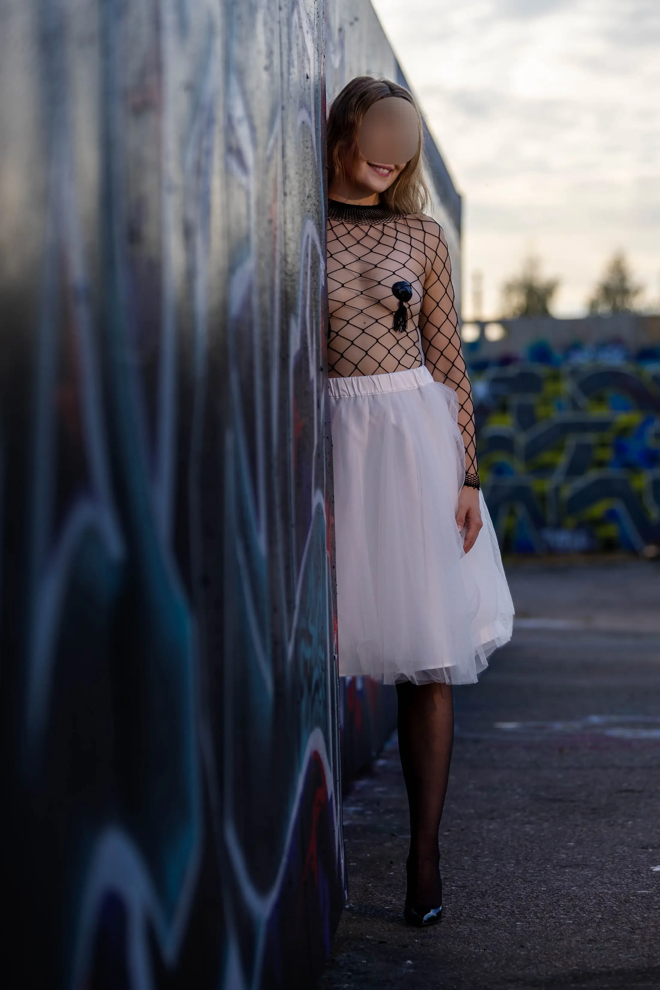 Josefine leans against a graffiti wall, dressed in a sheer black net top and white tulle skirt.