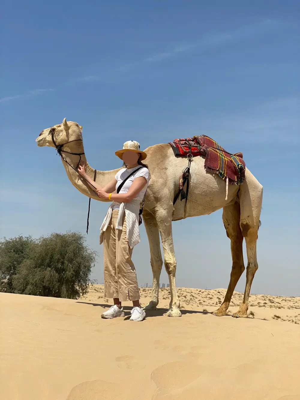Josefine in desert attire holds a camel against a clear sky in Dubai's sands.